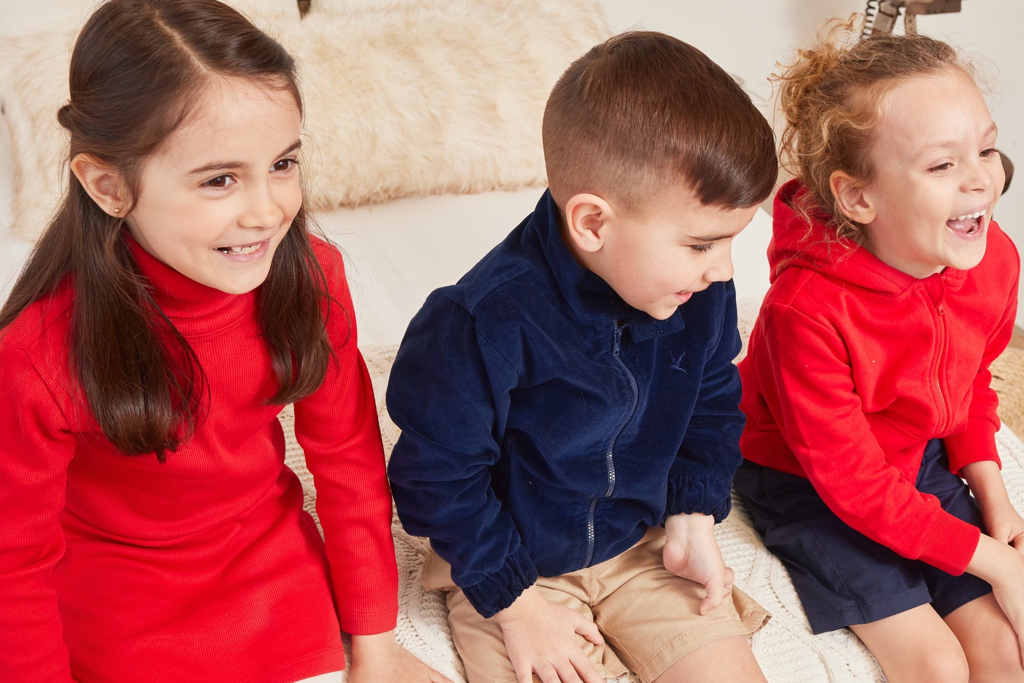 Three children sitting together on a bed, wearing red and blue clothing.